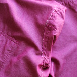 May include: Close-up of a pink fabric shirt with a pocket and a seam detail. The fabric is wrinkled and has a soft texture.