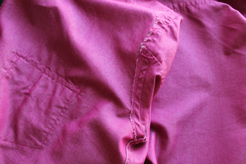 May include: Close-up of a pink fabric shirt with a pocket and a seam detail. The fabric is wrinkled and has a soft texture.