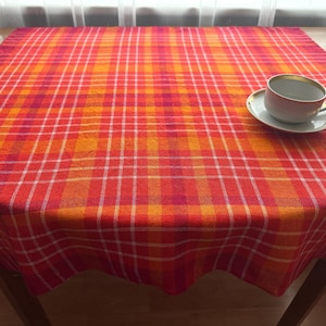 May include: A square tablecloth featuring a vibrant plaid pattern in shades of orange, red, and magenta with thin white intersecting lines. A white ceramic teacup with a gold rim sits on a matching saucer on the right side of the fabric.