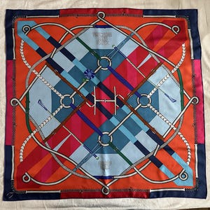 以下が含まれることがあります： 幾何学模様が特徴的な正方形のシルクスカーフ。ネイビーブルーの縁取りがあり、青、オレンジ、赤、ピンクの色合いで、交差するストラップ、鞭、リングのパターンが中央にあります。「HERMES」の文字が見えます。
