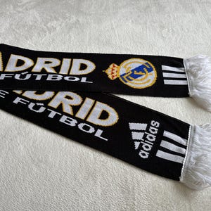 May include: Black scarf with white and gold text reading "MADRID F&Uacute;TBOL". Features the Real Madrid crest and Adidas logo. The scarf has white fringe on the ends.