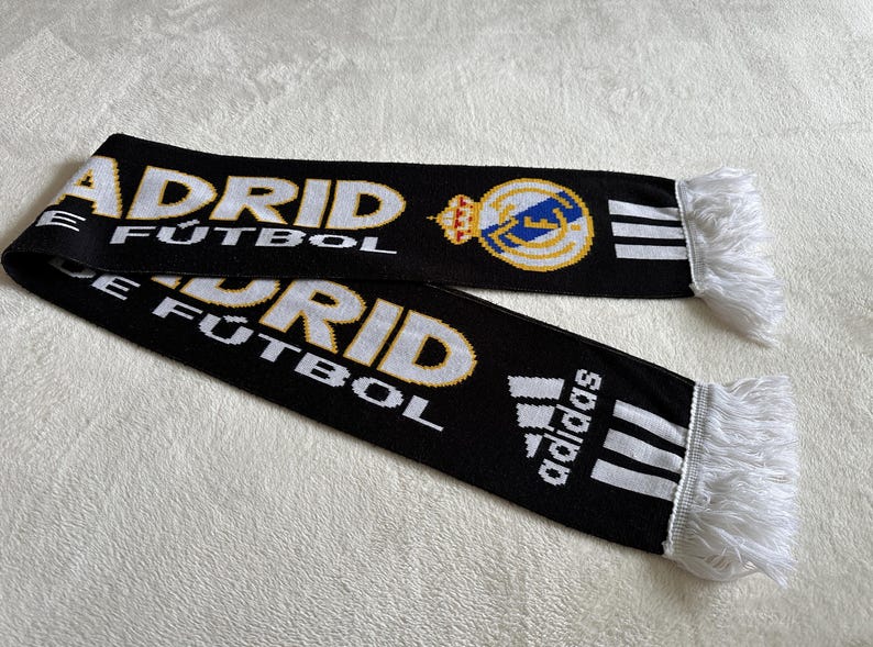 May include: Black scarf with white and gold text reading "MADRID F&Uacute;TBOL". Features the Real Madrid crest and Adidas logo. The scarf has white fringe on the ends.