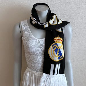 May include: Black and white knitted scarf with the Real Madrid crest. The scarf features the team's logo in yellow, blue, and white, with a crown detail. The scarf has white fringe at the bottom and is displayed on a mannequin.