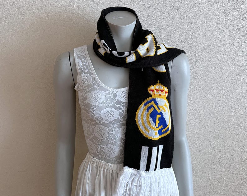 May include: Black and white knitted scarf with the Real Madrid crest. The scarf features the team's logo in yellow, blue, and white, with a crown detail. The scarf has white fringe at the bottom and is displayed on a mannequin.
