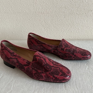 May include: A pair of red and black snakeskin-patterned leather loafers. The slip-on shoes have a rounded toe and a low heel. The shoes are displayed on a light-coloured surface.