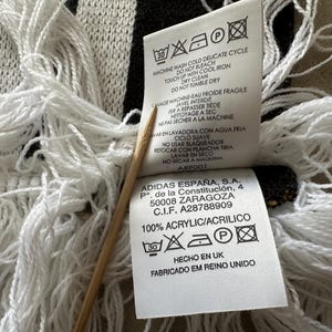 May include: Close-up of a black and white fringed scarf with care instructions and a tag that reads "100% ACRYLIC/ACRILICO" and "HECHO EN UK". The scarf has a woven pattern and long white fringe.