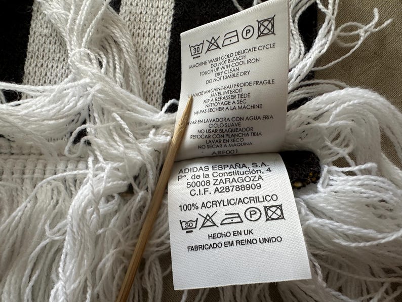 May include: Close-up of a black and white fringed scarf with care instructions and a tag that reads "100% ACRYLIC/ACRILICO" and "HECHO EN UK". The scarf has a woven pattern and long white fringe.