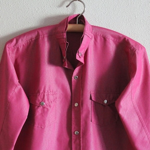 May include: A pink long-sleeved button-down shirt with two chest pockets and silver buttons.