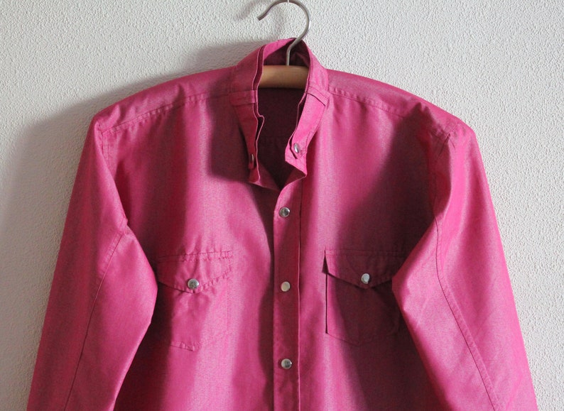 May include: A pink long-sleeved button-down shirt with two chest pockets and silver buttons.