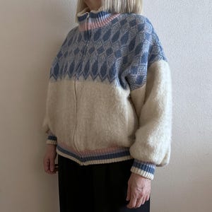 May include: A zip-up sweater with a blue diamond pattern on the upper portion and cream-colored lower section. The sweater has a high collar with pink and blue accents, and matching cuffs. The sleeves are puffy.