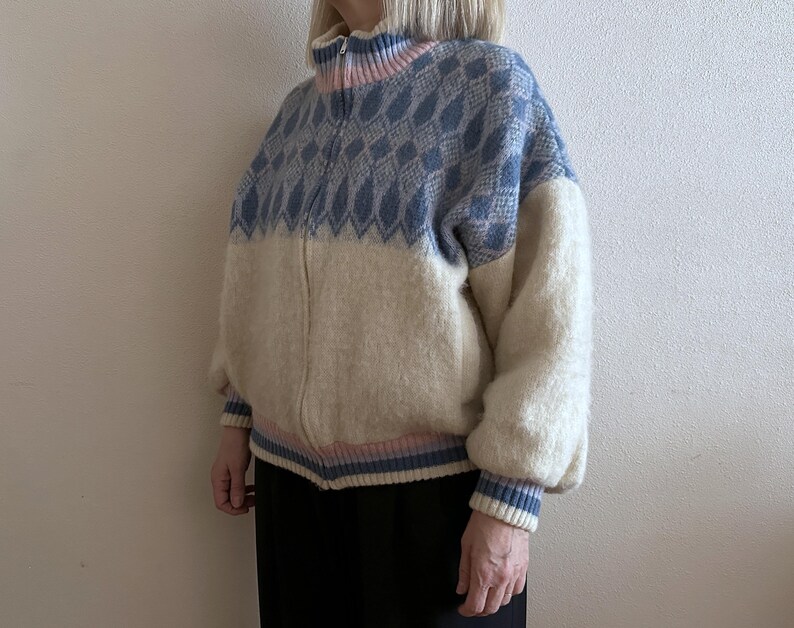 May include: A zip-up sweater with a blue diamond pattern on the upper portion and cream-colored lower section. The sweater has a high collar with pink and blue accents, and matching cuffs. The sleeves are puffy.
