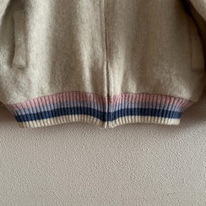 May include: A cream-colored, fuzzy jacket with a ribbed hem and cuffs. The hem and cuffs feature a striped design in pink, blue, and white. The jacket has a zipper closure and a soft, textured appearance, suitable for outerwear.