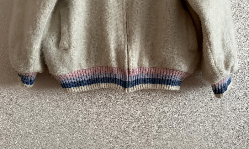 May include: A cream-colored, fuzzy jacket with a ribbed hem and cuffs. The hem and cuffs feature a striped design in pink, blue, and white. The jacket has a zipper closure and a soft, textured appearance, suitable for outerwear.