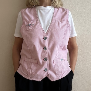 Può includere: Un gilet a righe rosa e bianche con scollo a V, chiusura con bottoni e due tasche frontali. Il gilet presenta cuciture decorative e piccoli dettagli ricamati. È indossato sopra una maglietta bianca e pantaloni neri.