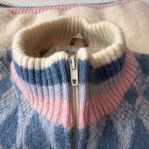May include: Close-up of a vintage zip-up sweater with a blue and white diamond pattern. The collar features stripes of white, blue, and pink. The sweater is made of a soft, fuzzy material.