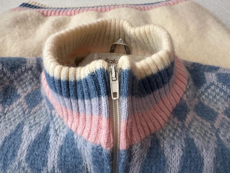 May include: Close-up of a vintage zip-up sweater with a blue and white diamond pattern. The collar features stripes of white, blue, and pink. The sweater is made of a soft, fuzzy material.