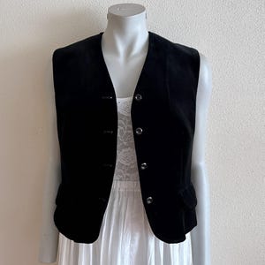 Può includere: Un gilet in velluto nero con scollo a V, quattro bottoni frontali e due piccole tasche con patta in vita. Il gilet è indossato sopra un top in pizzo bianco e una gonna bianca, presentando un design strutturato con una finitura scura.