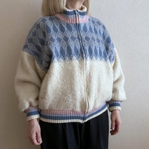 May include: A zip-up sweater with a high collar. The top half is light blue with a diamond pattern, and the bottom half is off-white. The collar, cuffs, and hem feature pink and blue stripes. The sweater is made of a soft, textured material.