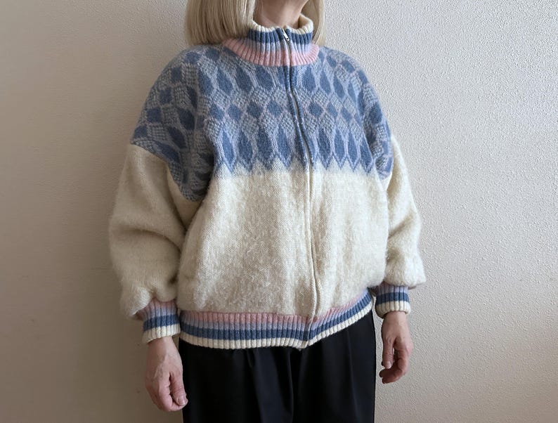 May include: A zip-up sweater with a high collar. The top half is light blue with a diamond pattern, and the bottom half is off-white. The collar, cuffs, and hem feature pink and blue stripes. The sweater is made of a soft, textured material.