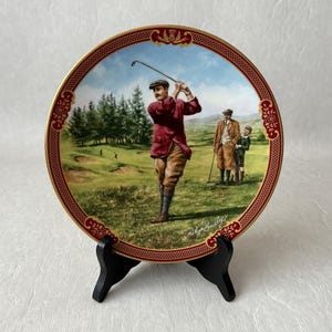May include: Decorative plate featuring a golf scene with a golfer in a red jacket, brown pants, and black socks. The plate has a gold rim with red accents and is displayed on a black stand. The background shows a golf course with trees and other golfers.
