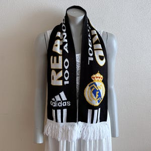 May include: Black and white knit scarf with the text "REAL 100 A&Ntilde;OS" and the Adidas logo. Features a team crest and white fringe. A sports accessory.