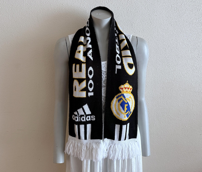 May include: Black and white knit scarf with the text "REAL 100 A&Ntilde;OS" and the Adidas logo. Features a team crest and white fringe. A sports accessory.