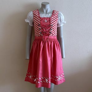 Può includere: Un abito di dirndl rosa e bianco con un corpetto a quadri rossi e bianchi e bordi bianchi. L'abito ha una sovrapposizione di pizzo bianco e un design a forma di cuore sul corpetto.