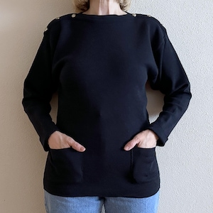 May include: A black long-sleeved top with a boat neck and two front pockets. The top has gold-colored buttons on the shoulders. The model is wearing blue jeans. This top is a casual style.