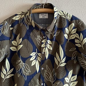Puede incluir: Una camisa de manga corta con estampado de hojas. La camisa tiene un fondo marrón con diseños de hojas azul marino y crema. El cuello tiene una etiqueta que dice "JEAN PASCALE XXL".