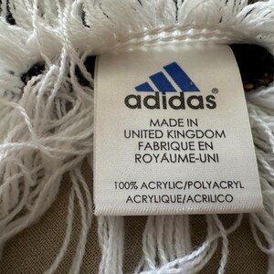 May include: Close-up of a white fringed textile with a tag. The tag features the Adidas logo and text indicating it's made in the United Kingdom from 100% acrylic. The fringe is thick and textured.