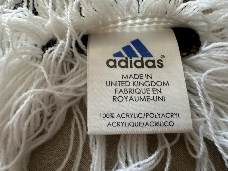 May include: Close-up of a white fringed textile with a tag. The tag features the Adidas logo and text indicating it's made in the United Kingdom from 100% acrylic. The fringe is thick and textured.