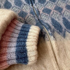 May include: Close-up of a cozy, textured sweater with a blue and white diamond pattern. The sleeve cuff features blue and white stripes. The zipper is visible, and the overall color palette is soft and inviting.