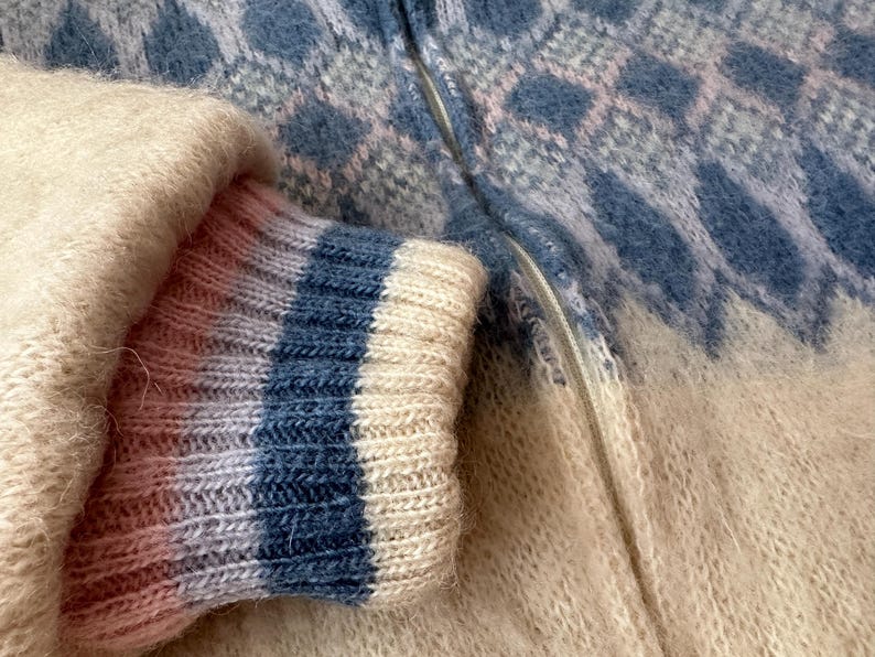 May include: Close-up of a cozy, textured sweater with a blue and white diamond pattern. The sleeve cuff features blue and white stripes. The zipper is visible, and the overall color palette is soft and inviting.