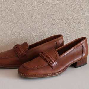 Könnte beinhalten: Ein Paar braune Lederschuhe mit einem geflochtenen Detail an der Vorderseite. Die Schuhe haben einen niedrigen Absatz und eine abgerundete Spitze.