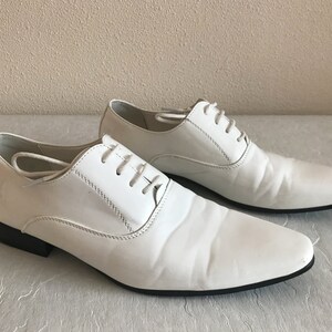 May include: Two pairs of white leather dress shoes with lace-up fastenings. The shoes have a pointed toe and a black rubber sole.