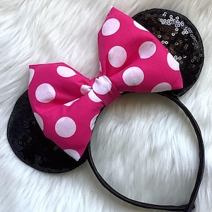 May include: A black headband with sequined ears and a large pink bow with white polka dots. The bow is centered on the headband, and the ears are on either side. The headband is on a white, fluffy surface.