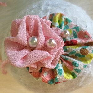 May include: A pink fabric flower with two white pearl embellishments, centered on a white background with a floral fabric underneath.