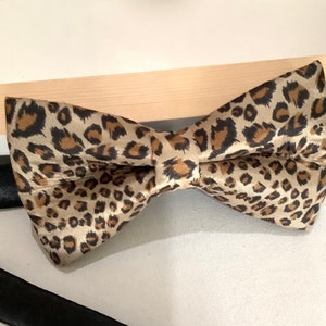 May include: A leopard print bow tie. The bow tie features a tan background with brown spots. The bow tie is made of a silky material. A black tie is visible in the background.