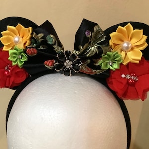 May include: Black headband with large black fabric ears decorated with red, yellow, and green fabric flowers and a black and gold bow with a jeweled center.