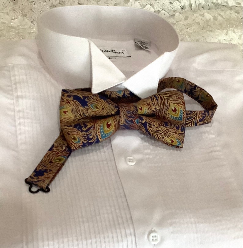 Puede incluir: Una pajarita con un estampado de plumas de pavo real en tonos dorados, azules y rojos. La pajarita est&aacute; sobre una camisa blanca con el frente plisado.