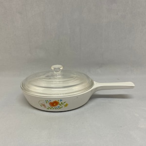 May include: A white saucepan with a clear glass lid and a long handle. The pan features a floral design with orange and blue flowers. The handle is white, and the lid has a clear knob.