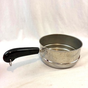 May include: A stainless steel saucepan with a black handle. The pan has a round shape with a perforated interior and a small loop at the end of the handle. The pan is sitting on a white surface.