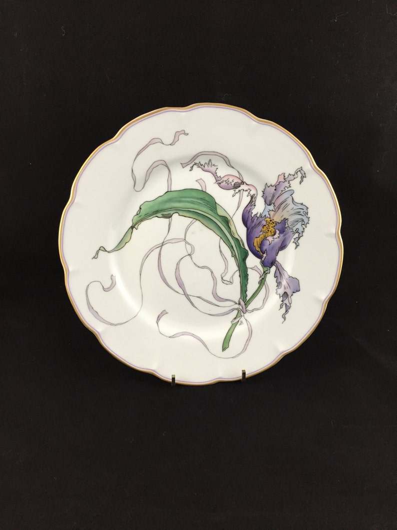 Peut inclure: Une assiette en porcelaine blanche avec un bord dor&eacute; et une fleur violette avec des feuilles vertes peintes dessus.