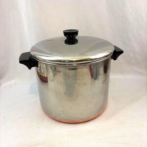 May include: A stainless steel cooking pot with a lid and black handles. The pot has a copper-colored bottom and a black knob on the lid. The pot is designed for cooking and food preparation.