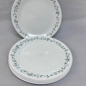 May include: A stack of white ceramic plates with a delicate floral design. The plates feature a border of green vines and small purple flowers. The plates are round and appear to be new.