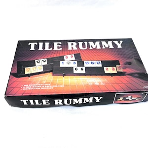 May include: A Tile Rummy game box with the game title in large white letters. The box features a sunset-like background and includes game tiles with numbers. The box also lists the contents: 106 engraved tiles, 4 tier racks, rack holders, and an instruction booklet.