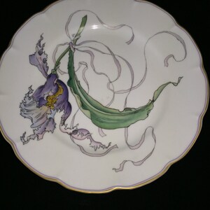 Peut inclure: Une assiette en porcelaine blanche avec un bord dor&eacute; pr&eacute;sente un motif peint &agrave; la main d'une orchid&eacute;e violette avec des feuilles vertes et des rubans roses.