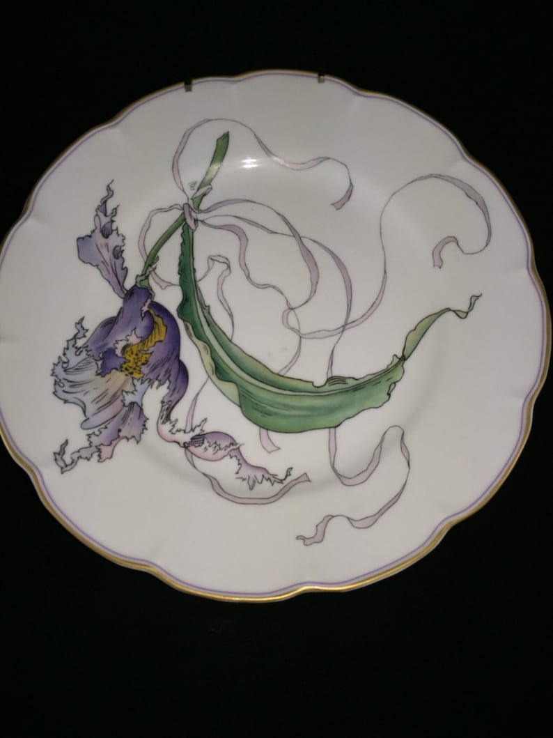 Peut inclure: Une assiette en porcelaine blanche avec un bord dor&eacute; pr&eacute;sente un motif peint &agrave; la main d'une orchid&eacute;e violette avec des feuilles vertes et des rubans roses.