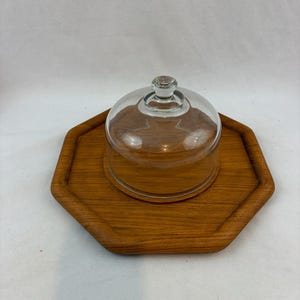 May include: A wooden serving tray with an octagonal shape and a clear glass dome. The tray is made of a light brown wood and has a slightly raised edge. The glass dome has a small knob on top.