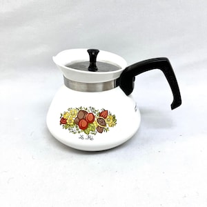 May include: White enamel tea kettle with a black handle and lid. The kettle features a decorative floral design with the words "Le Thé" printed on the front. The kettle has a stainless steel band around the middle.